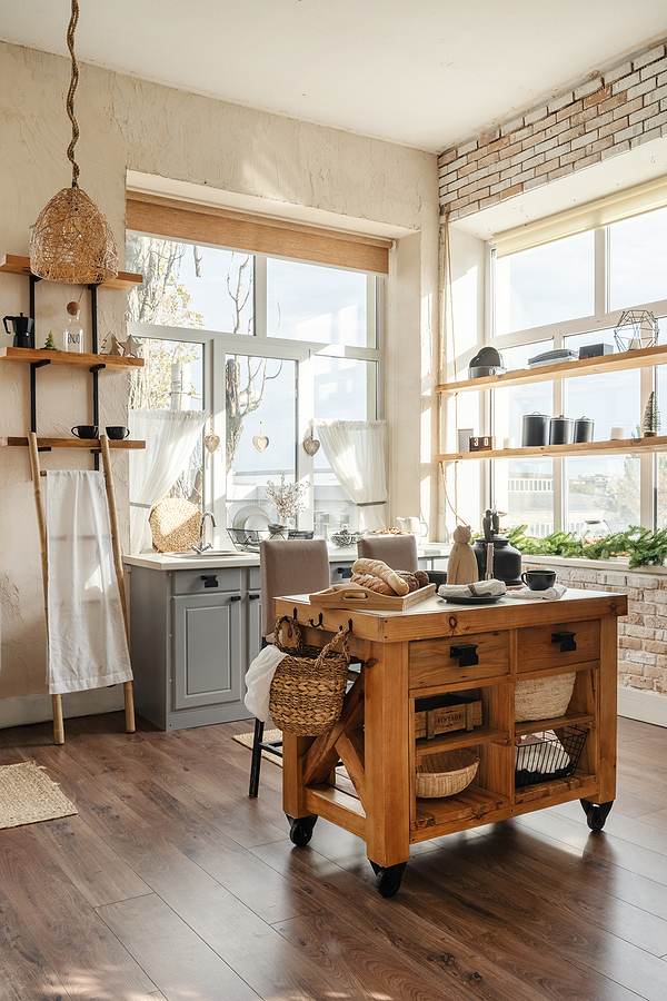 Vertical side view of stylish sunny kitchen interior with kitchenware, home decor and accessories, wooden table with homemade pastry food against cupboards and windows.